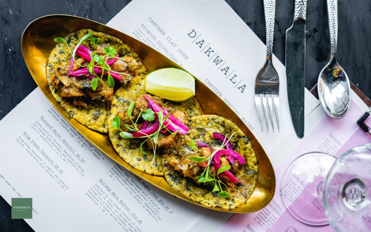 Three Indian-style tacos on a long gold plate surrounded by cutlery, a wine glass and a menu